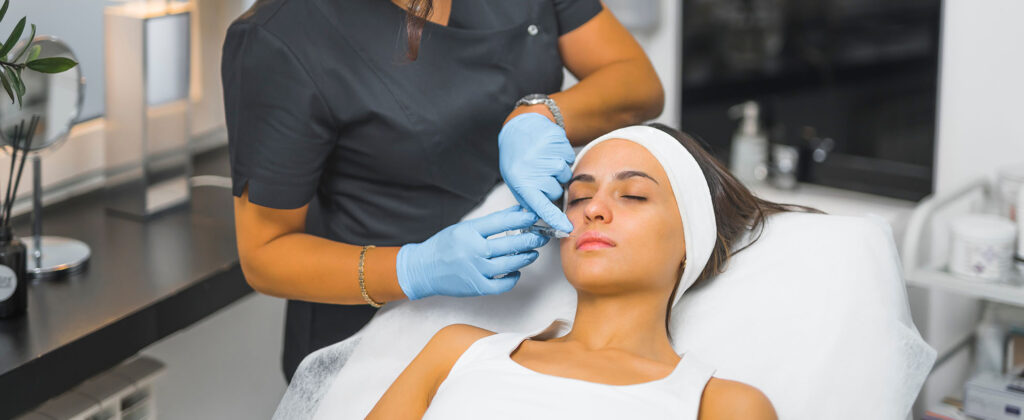 person lying on a treatment bed while an esthetician wearing blue gloves administers an injection near the lips