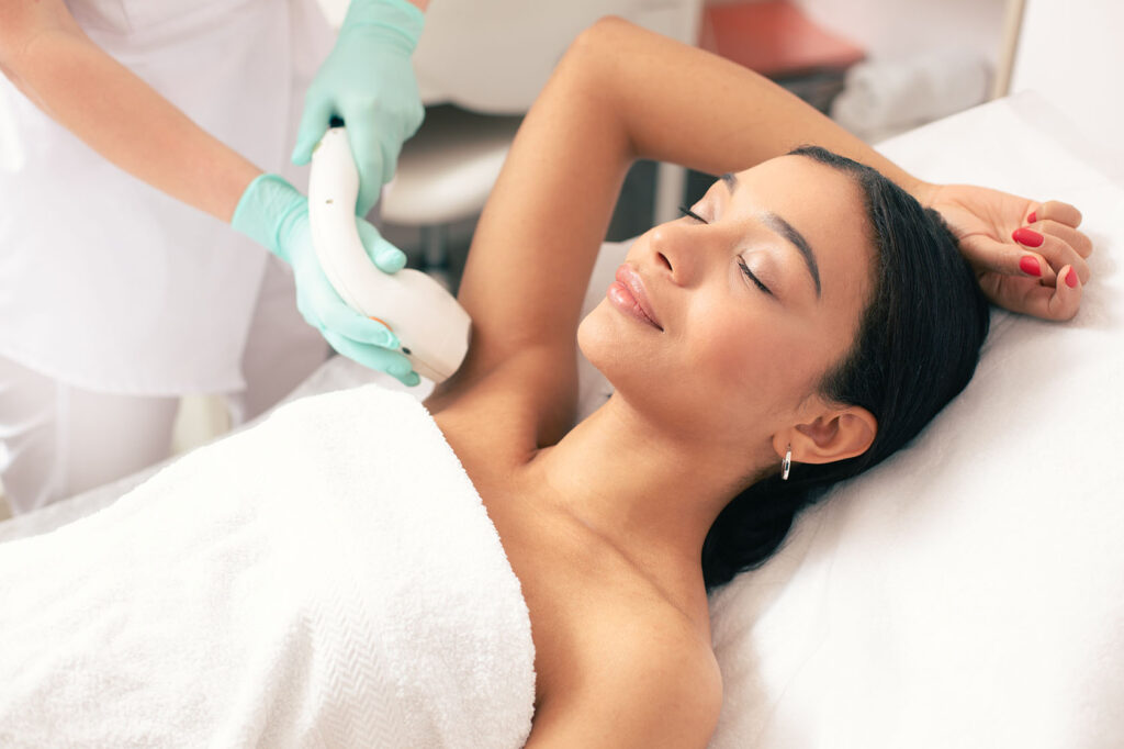 woman lying on treatment bed with towel while technician uses handheld laser device on her underarm