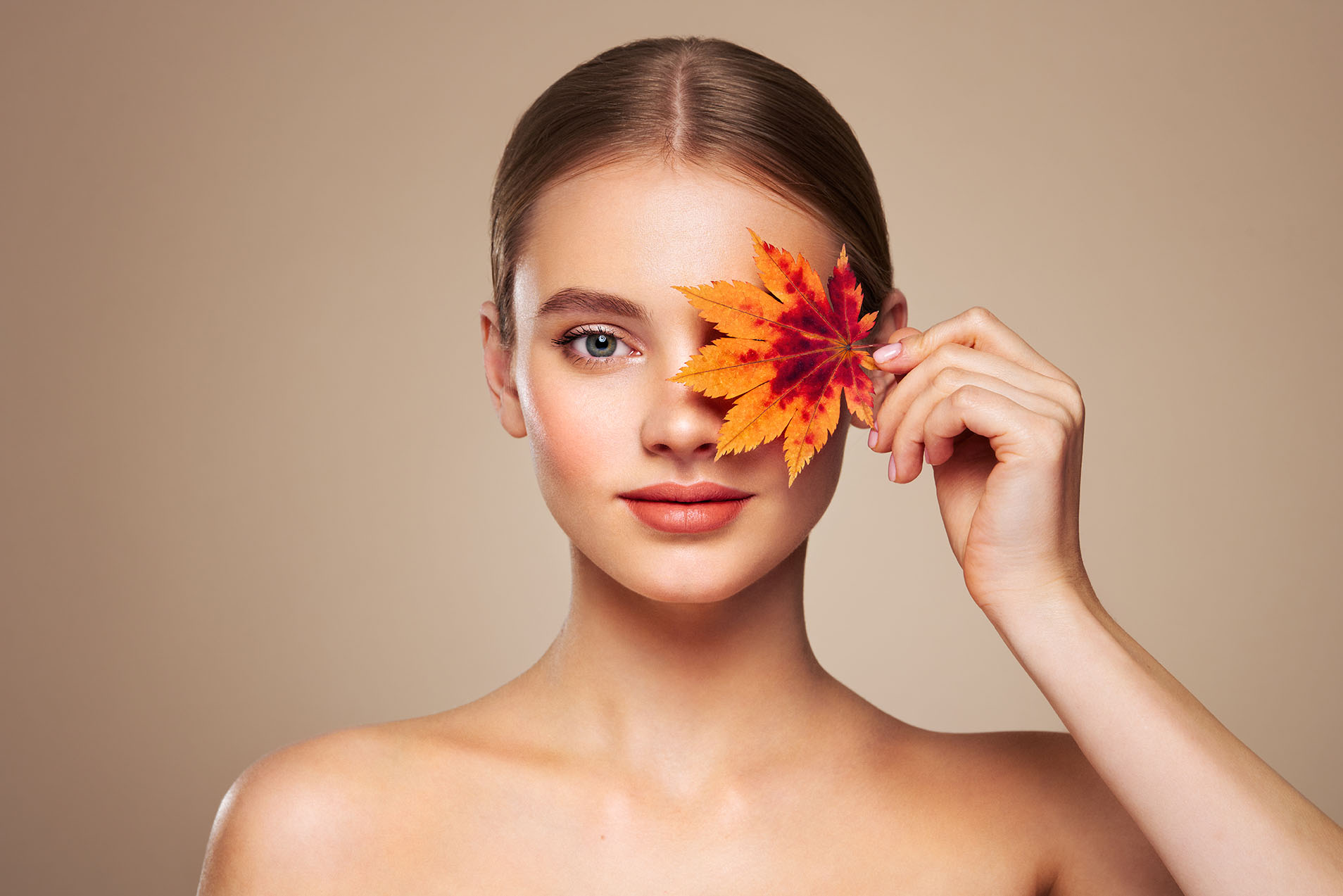 woman holding an orange maple leaf over one eye while standing against a neutral beige background