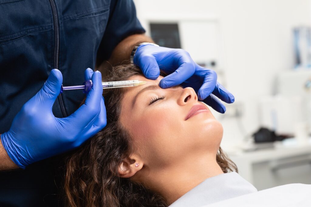 Person lying on a treatment chair while a practitioner wearing blue gloves positions a syringe near the forehead.