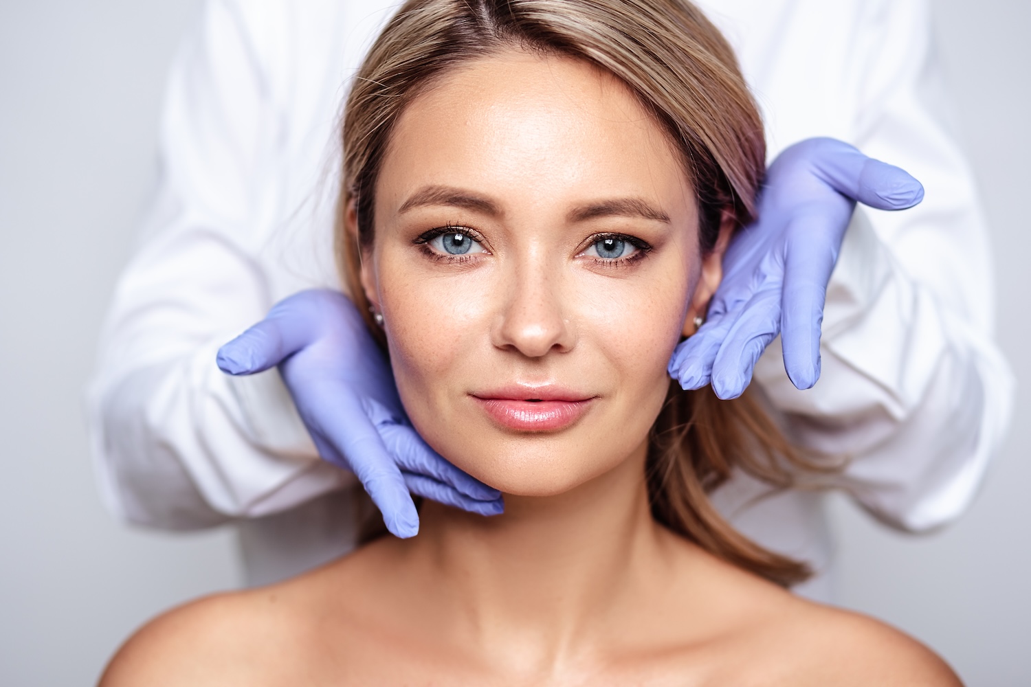Front-facing woman looking at the camera while a practitioner wearing gloves gently positions hands along her jaw and cheeks.