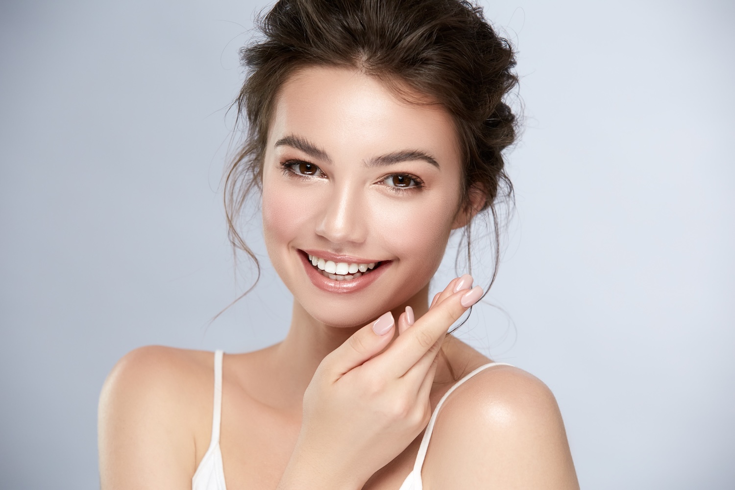 Woman smiling at the camera with hair pulled back, natural makeup, and one hand resting near her cheek against a light background.