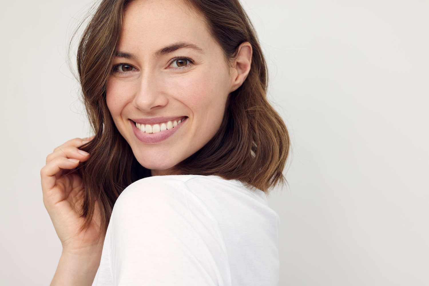 Woman smiling at the camera with shoulder-length brown hair, wearing a white top against a plain light background.