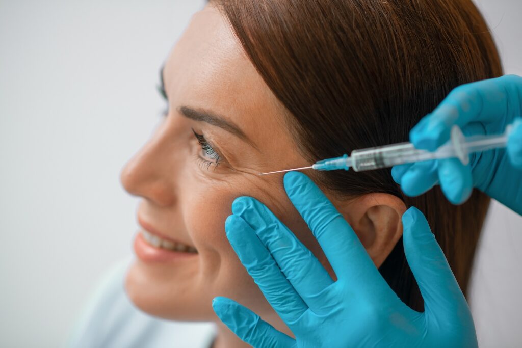 Side profile of a woman as a gloved practitioner holds her cheek and positions a syringe near the outer eye area.