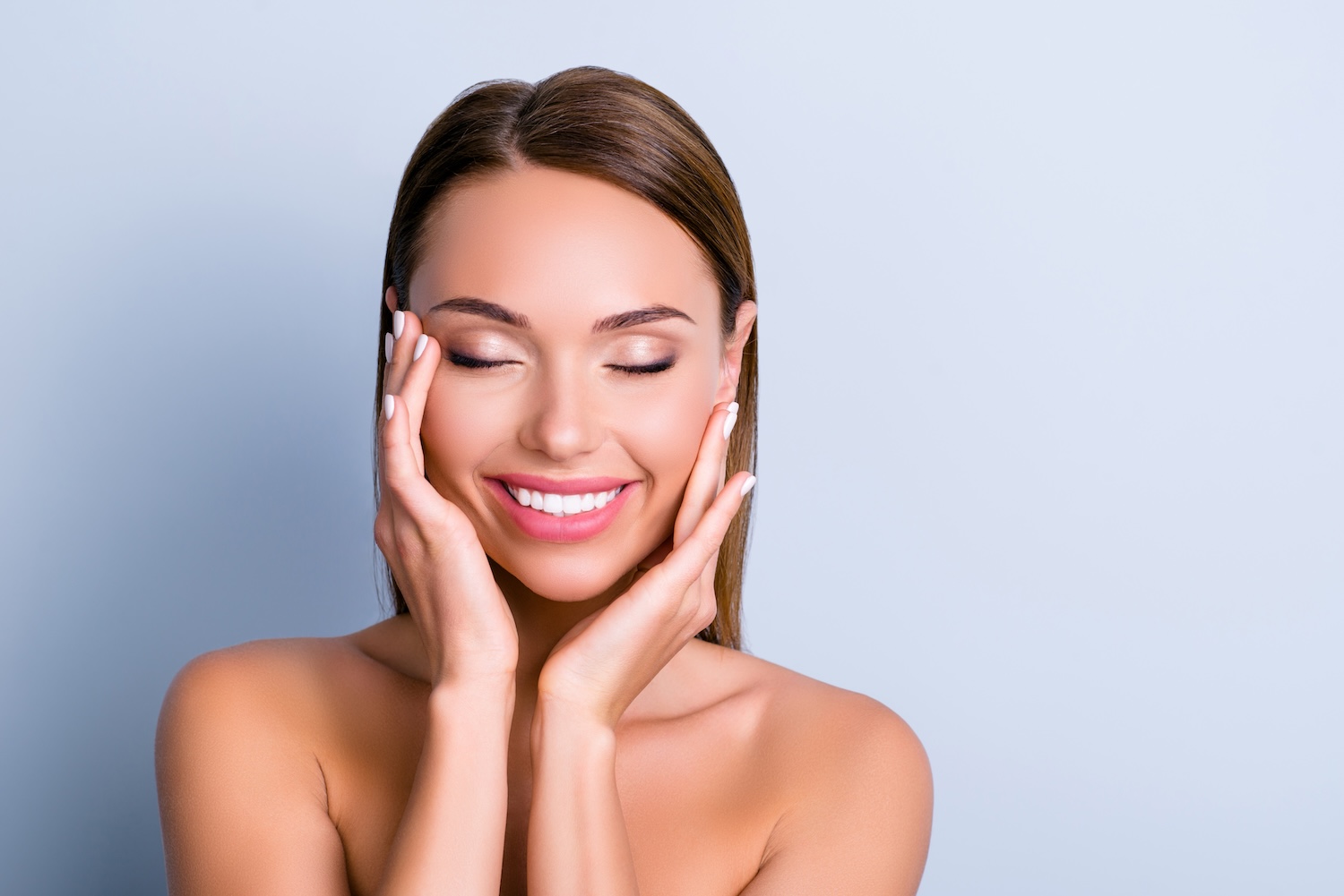 Woman with eyes closed smiling, both hands resting on her cheeks, against a plain light background.