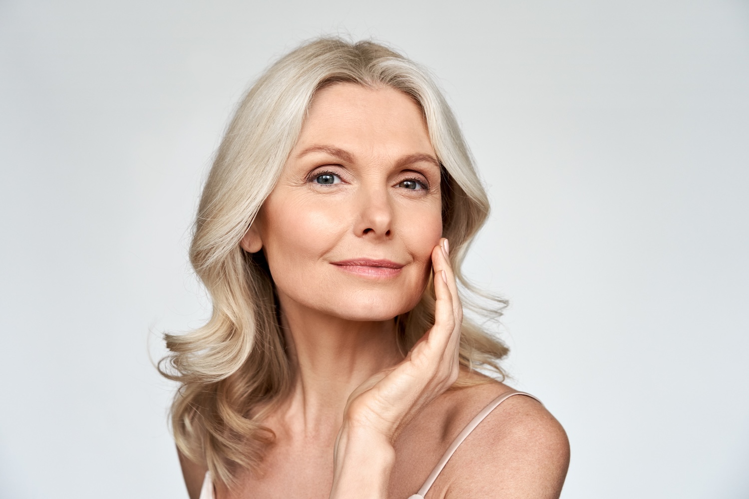 Woman with light hair looking at the camera, one hand resting on her cheek, against a plain light background.