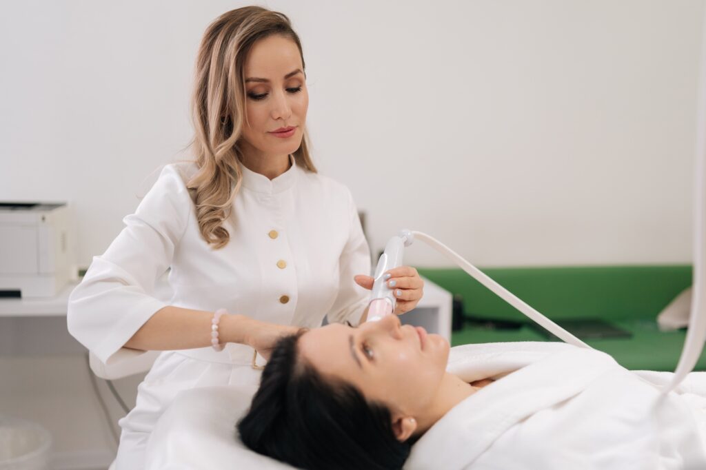 Practitioner in white uniform using a handheld device on a reclining patient’s face in a clinical setting.