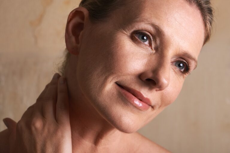Close-up of a woman’s face and neck with hand resting near collarbone against a neutral background.