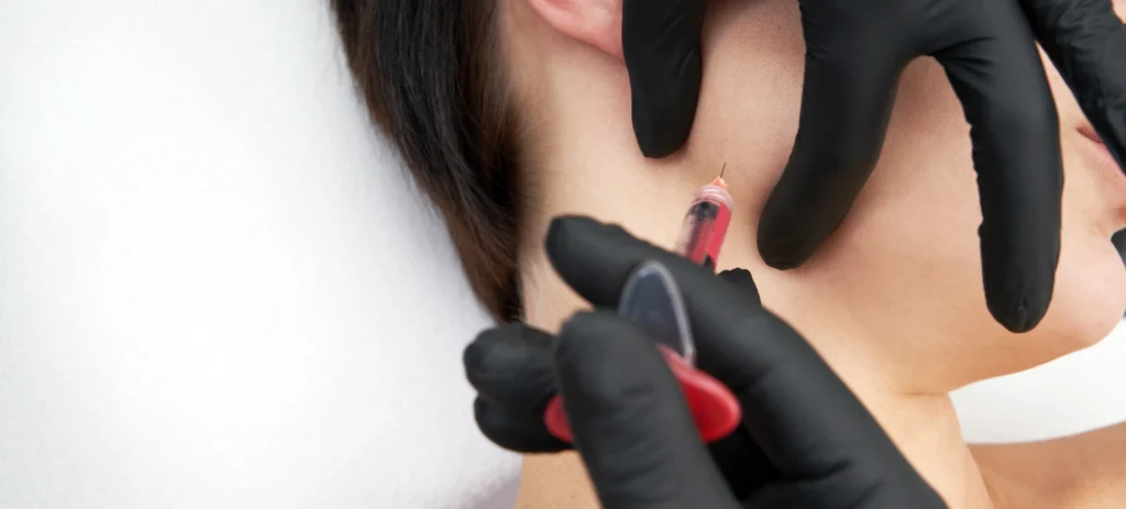 Gloved hands positioning a syringe near a woman's jawline during a facial injectable procedure