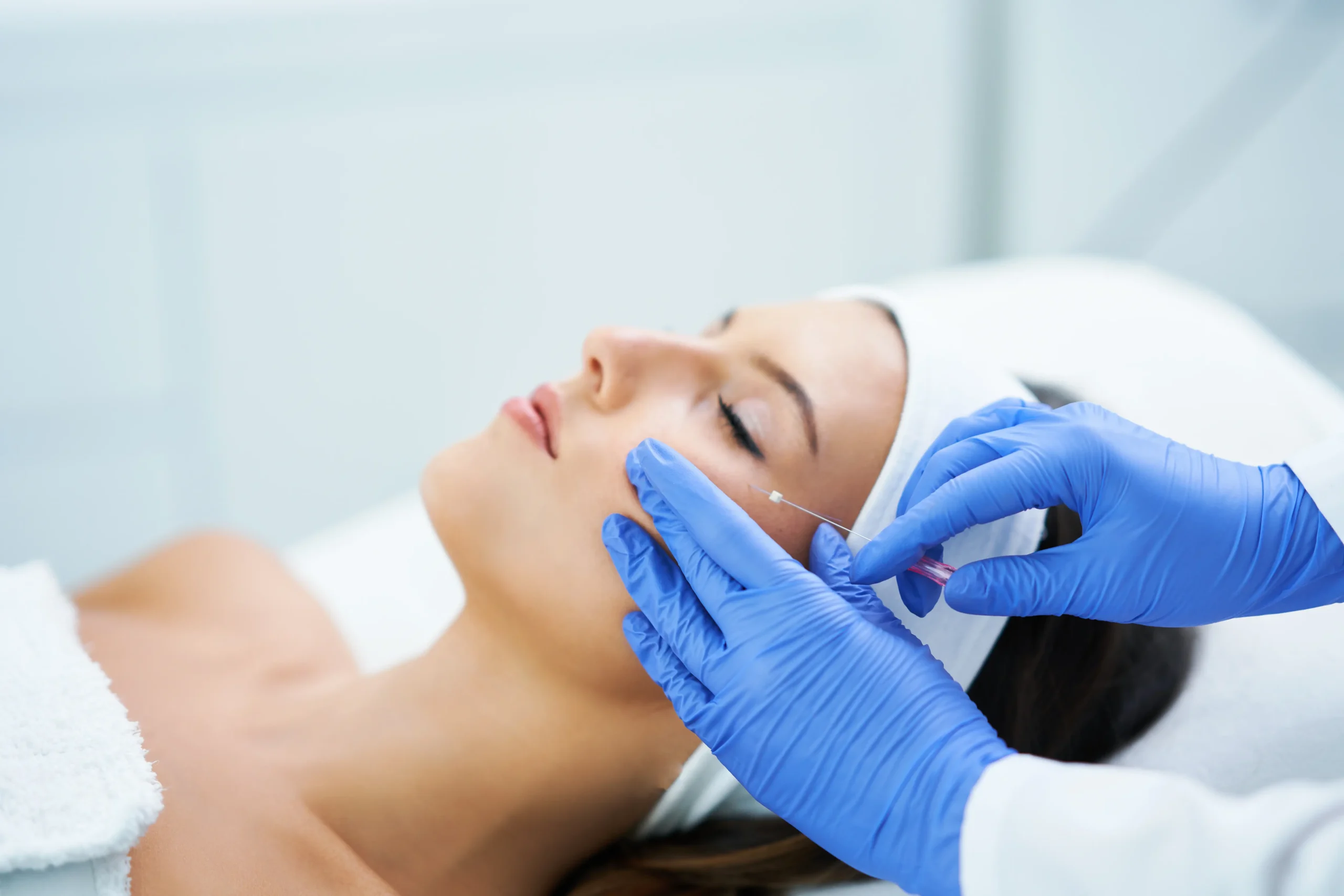 Gloved hands administering an injectable treatment to a woman's cheek while she reclines on a treatment bed
