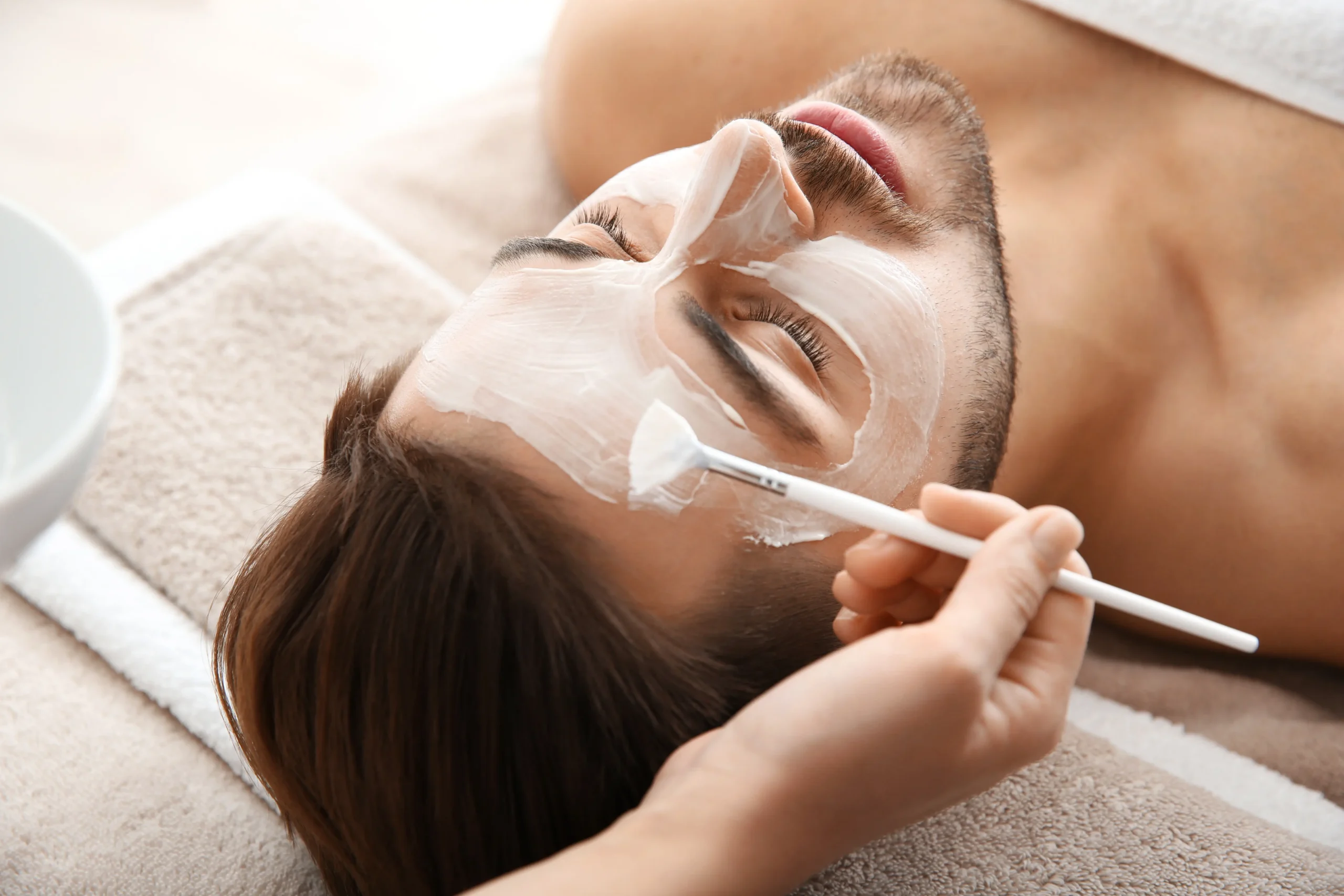 Practitioner applying a facial mask to a reclining man's face with a brush