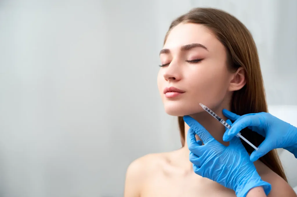 Gloved practitioner supports a woman's chin while positioning a syringe near her lower cheek and jawline.