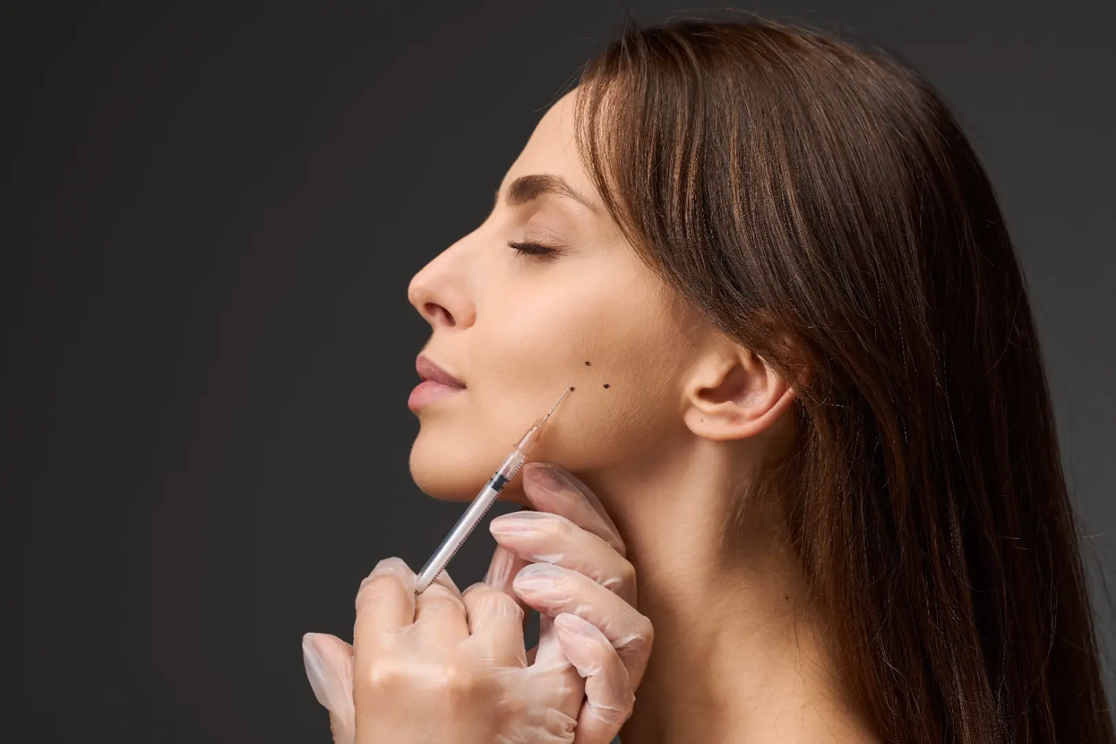 Gloved practitioner positions a syringe near marked points along a woman's lower cheek and jaw.