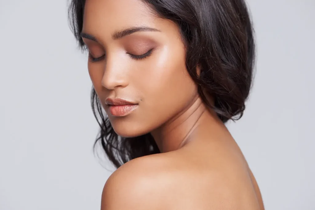 Dark-haired woman with closed eyes and a bare shoulder against a gray background.