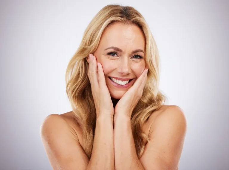 Smiling blonde woman rests both hands against her cheeks while facing the camera against a light background.