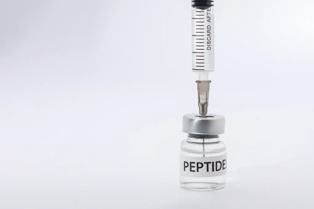 Syringe needle inserted through the cap of a clear vial labeled PEPTIDES against a white background.
