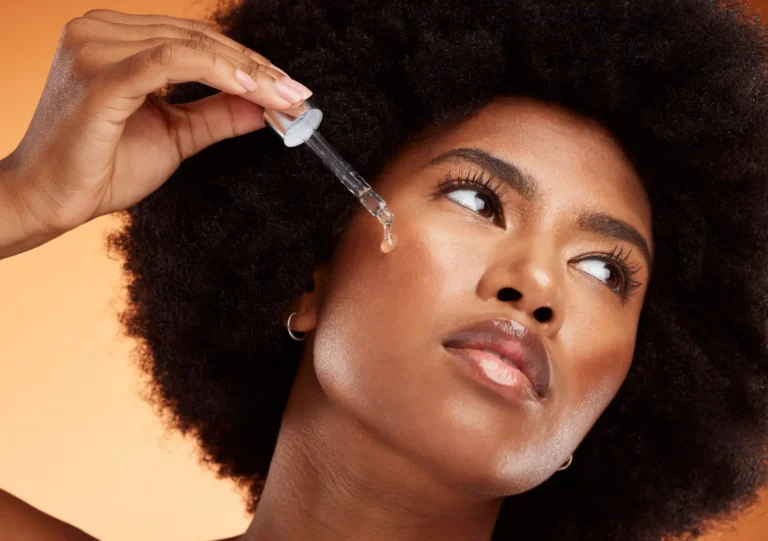 Woman with curly hair holding a glass dropper beside her cheek, with a clear droplet at its tip.