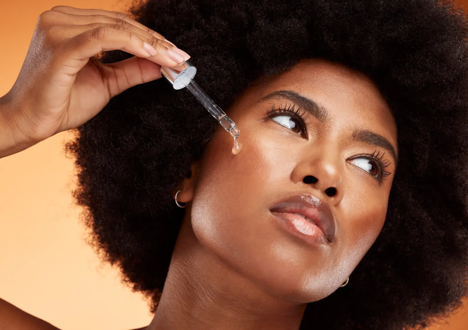 Woman with curly hair holding a glass dropper beside her cheek, with a clear droplet at its tip.
