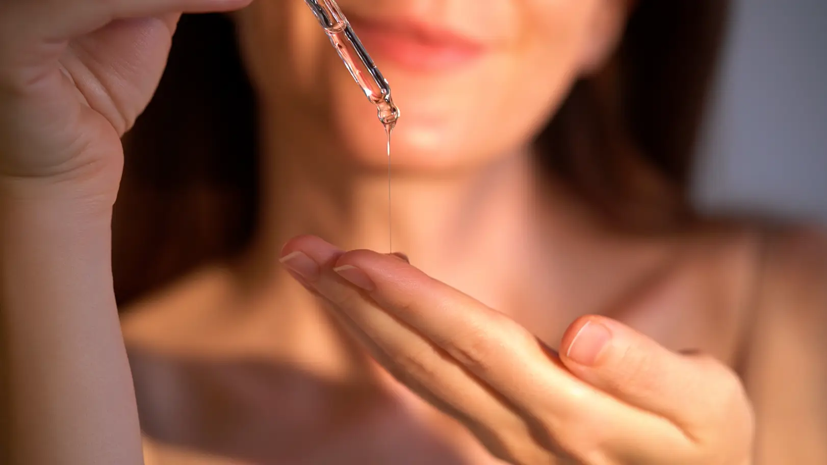 Clear serum drips from a glass dropper onto a person’s fingertips in warm light.