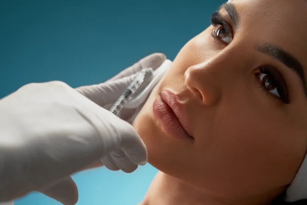 Gloved hands position a syringe beside a woman’s cheek as she lies against a blue background.